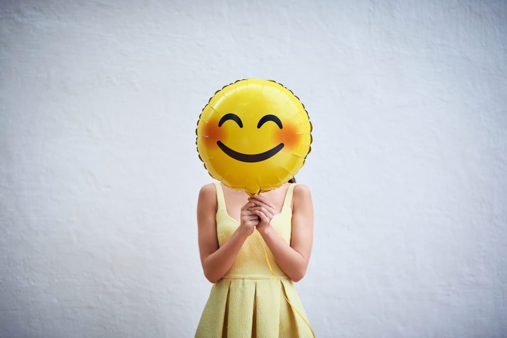 A woman holding a smiling emoji balloon covering her face.